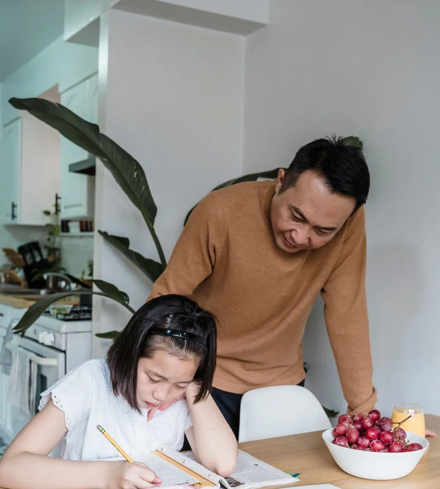 Người bố cúi xuống xem con gái làm bài tập ở bàn ăn